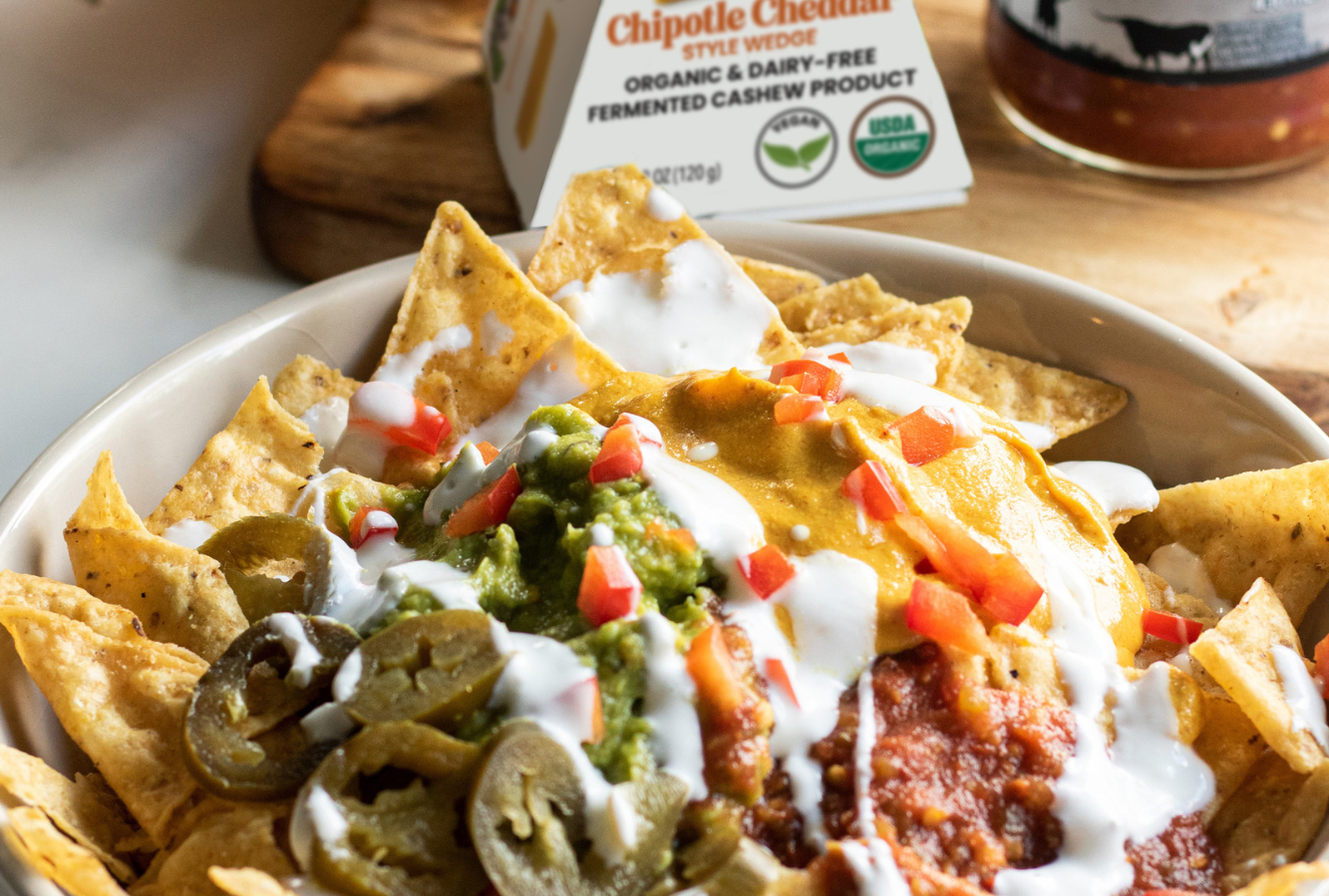 Hand scooping a tortilla chip from a bowl of nachos topped with guacamole, salsa, and vegan chipotle cheddar sauce. Served next to a bag of tortilla chips and a box of vegan chipotle cheddar cheese.