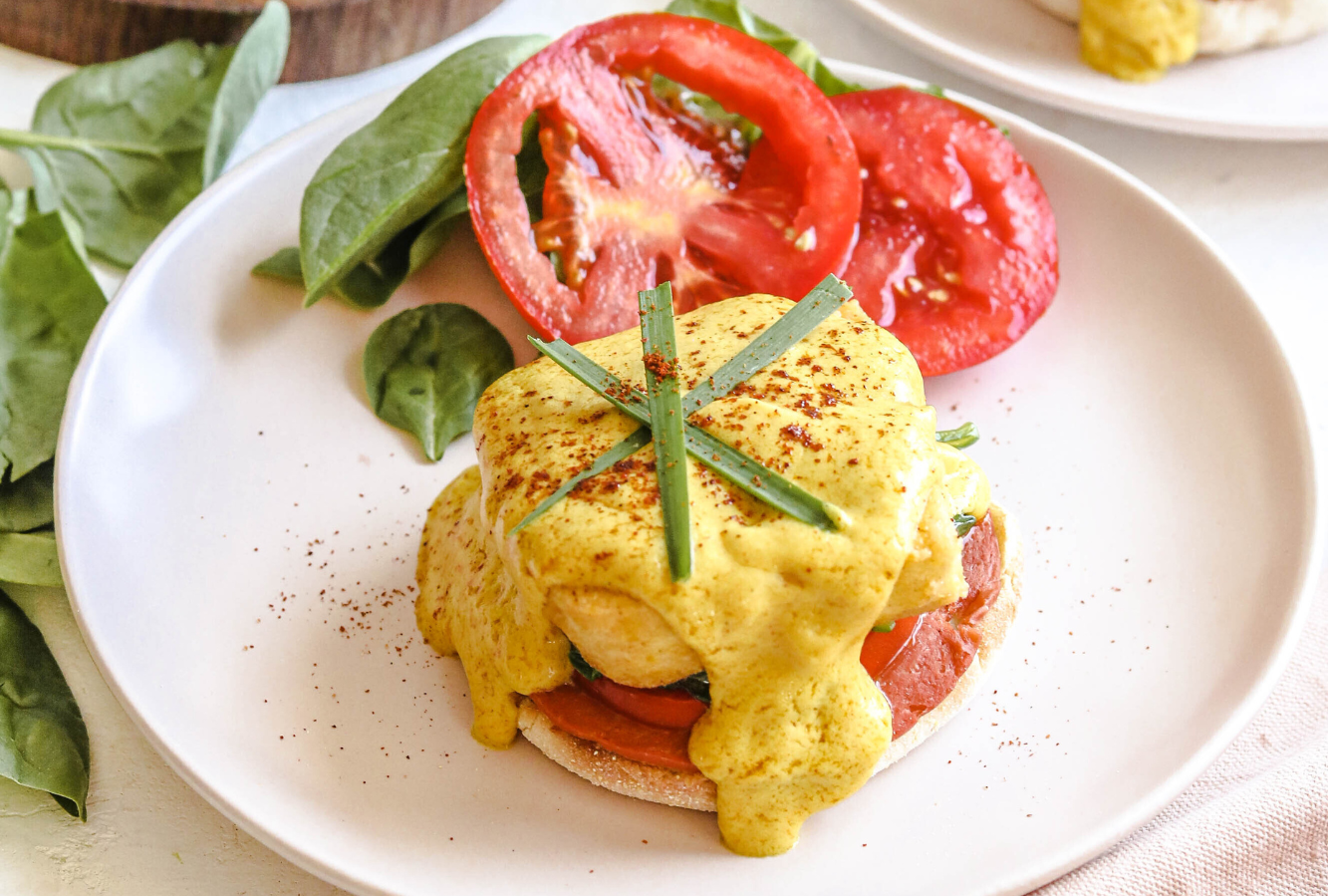 A plate with English muffins prepared with vegan eggs and a dairy-free hollandaise sauce. Served with fresh green salad next to a box of dairy-free smoky gouda cheese.