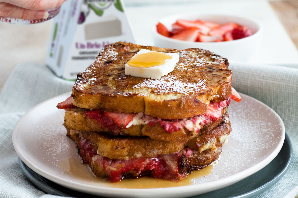 A stack of French toast on a plate, drizzled with maple syrup and stuffed with strawberries and dairy-free brie cheese. A spoon is adding a dollop of strawberry compote to the top and a box of dairy-free brie cheese is in the background.