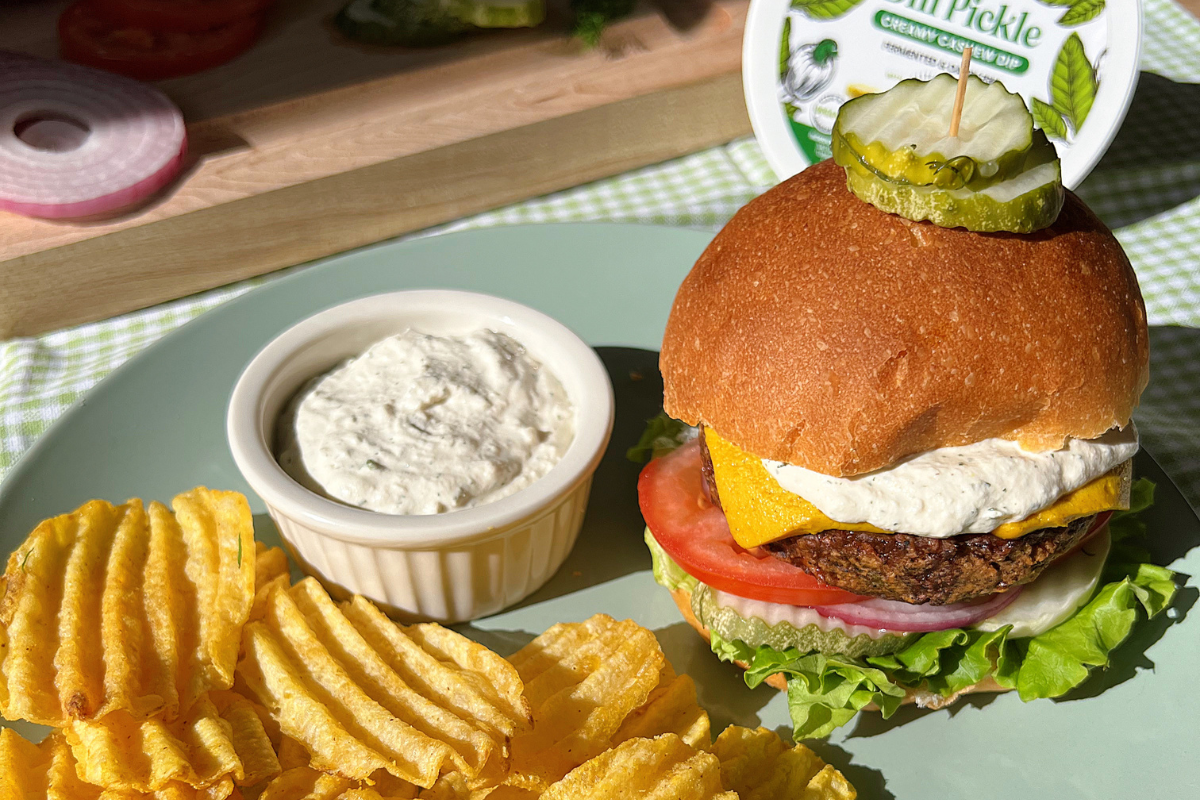 Veggie burger with Nuts For Cheese dill pickle dip, sharp cheddar style cheese and an extra side of dip with ruffle chips on a plate.