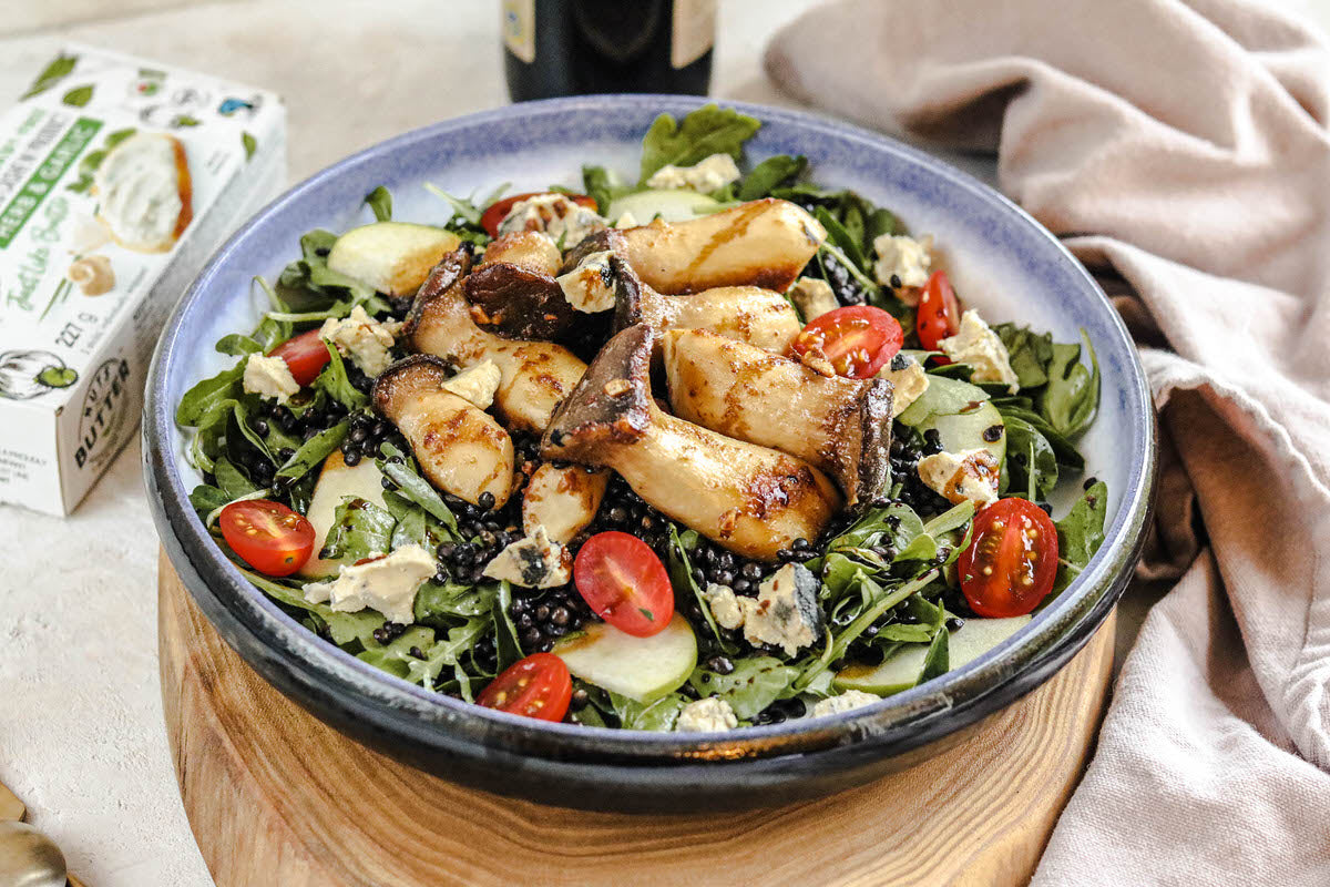 Bowl of salad topped with roasted mushrooms, black lentils, and chunks of dairy-free black garlic cheese.