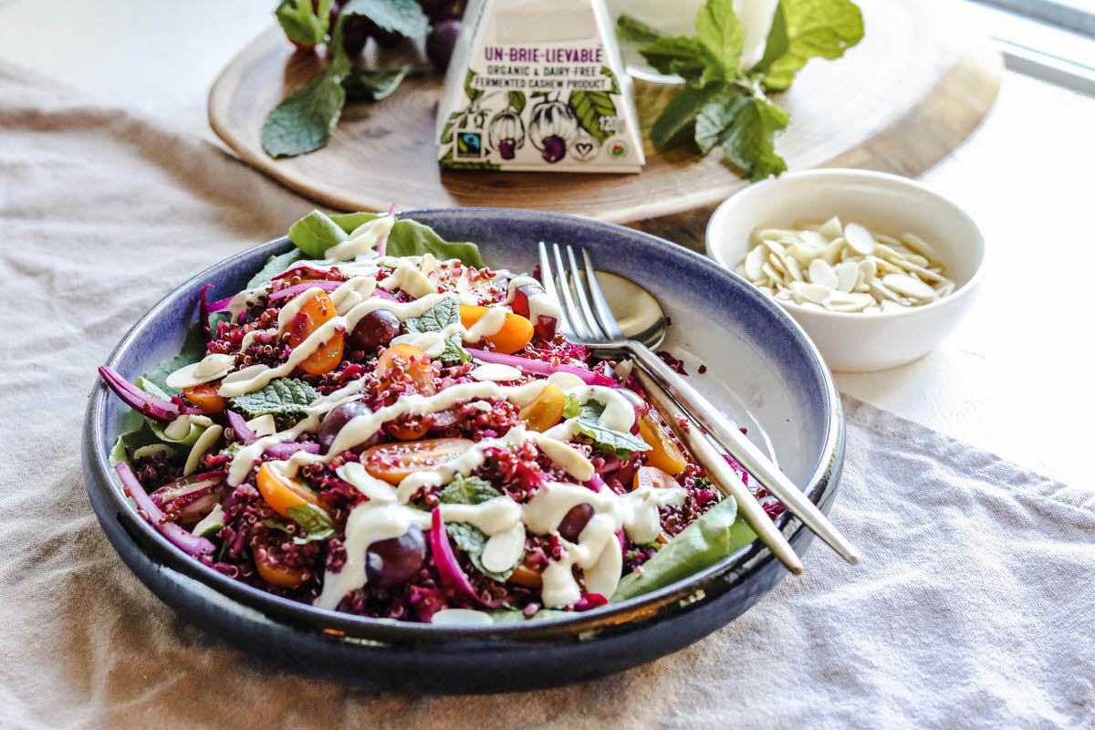 Bowl of salad topped with red quinoa, fresh tomatoes, red onions, and a drizzle of dairy-free dressing. Served next to a bowl of slivered almonds and a box of dairy-free brie cheese.