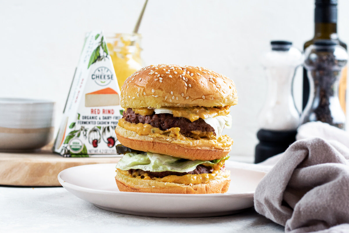 A vegan big-mac made with dairy-free cheese sauce sits on a plate. In the background is a box of dairy-free smoked gouda cheese.