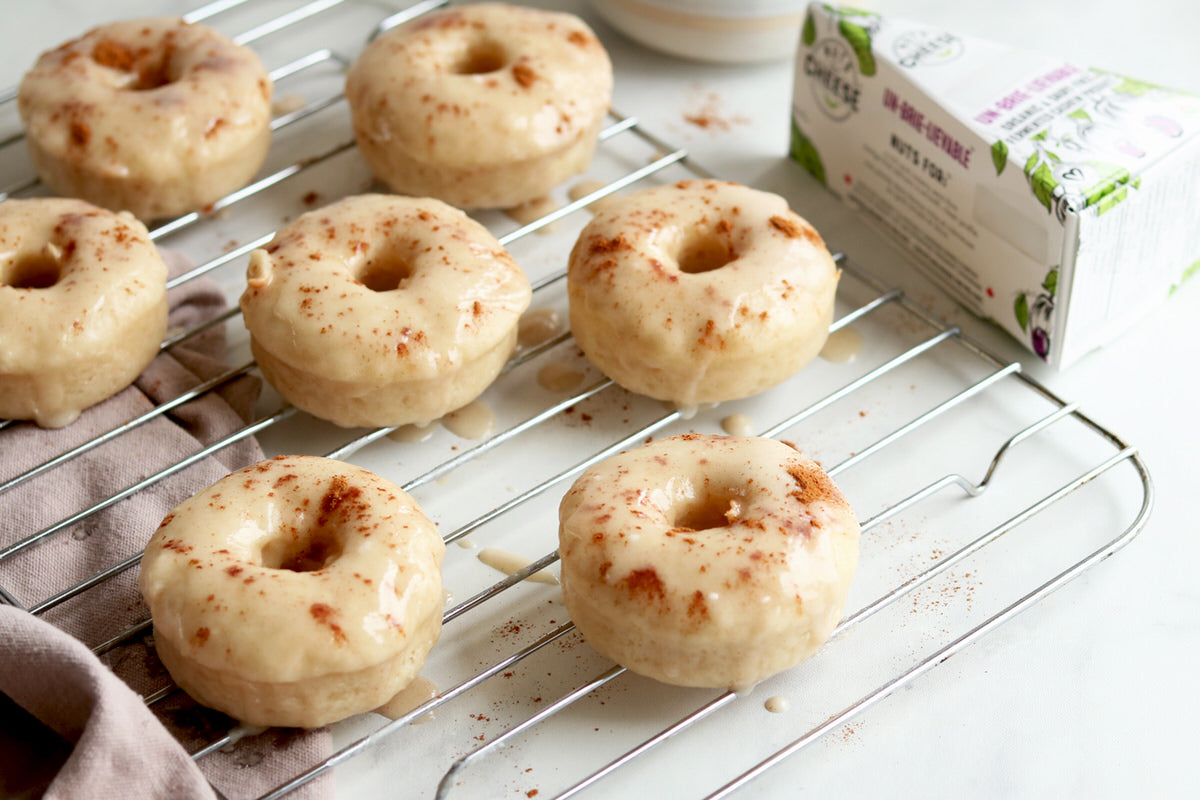 Baked donuts on a cooling rack, covered in a dairy-free brie and cinnamon glaze set beside a box of dairy-free brie cheese.