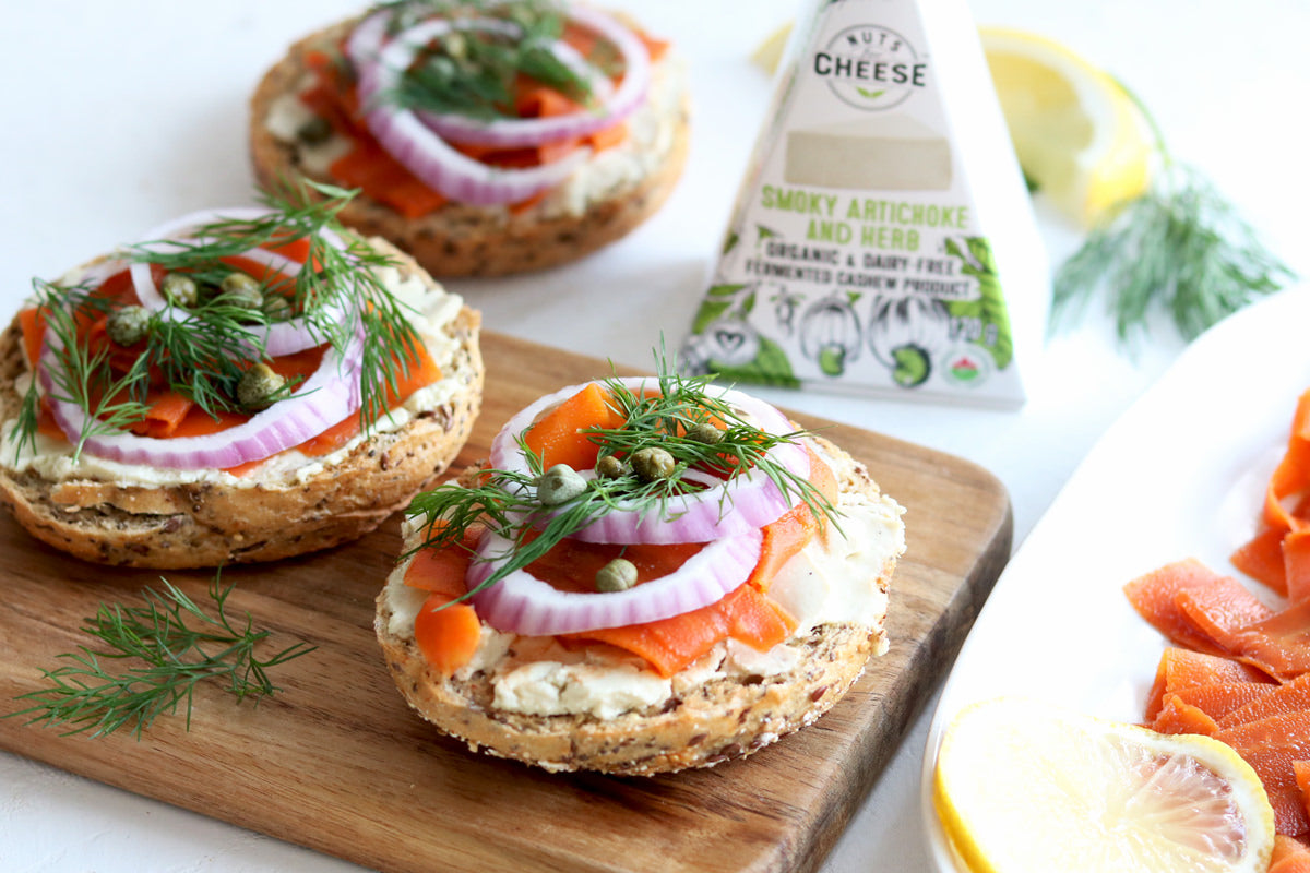 Three open-faced bagels topped with dairy-free artichoke & herb cheese, vegan carrot lox, red onions, capers, and fresh dill. Served next to a box of dairy-free artichoke & herb cheese.