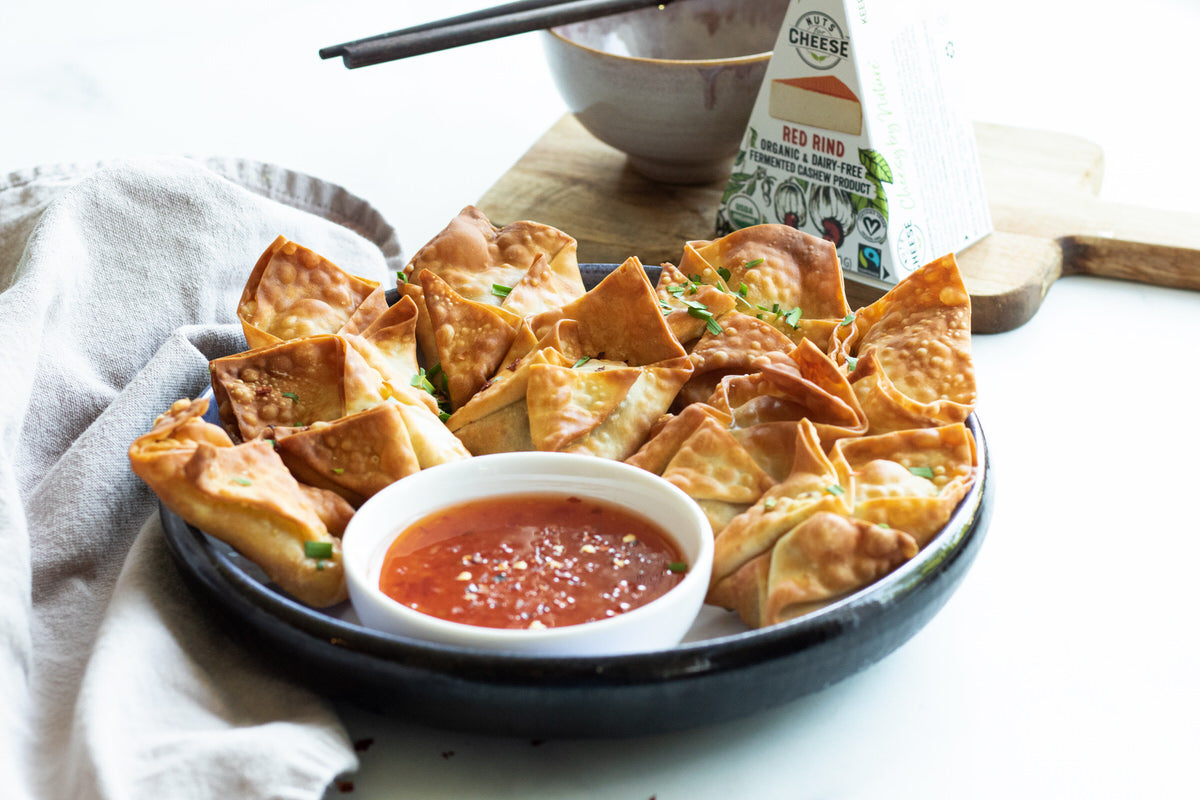 Large bowl of air fried vegan wontons stuffed with dairy-free gouda cheese and served with a bowl of sweet chili sauce. Box of dairy-free smoked gouda cheese in the background.