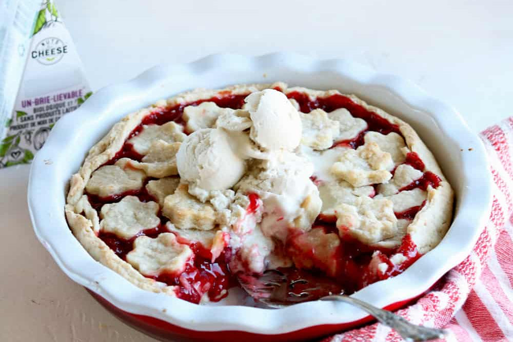 Baked cherry pie topped with scoops of dairy-free brie ice cream and served next to a box of dairy-free brie cheese.