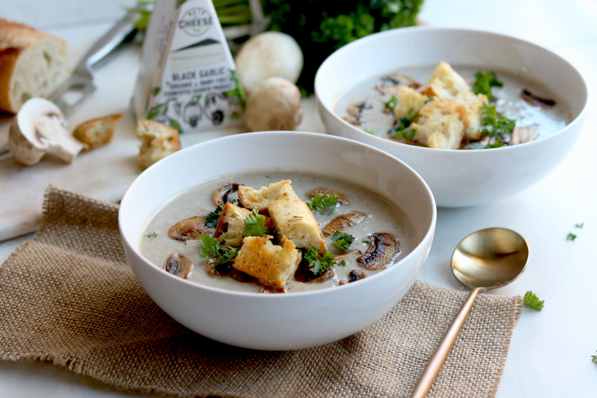 Two bowls of creamy vegan mushroom soup made with dairy-free black garlic cheese and topped with toasted croutons. Served next to a box of dairy-free black garlic cheese.