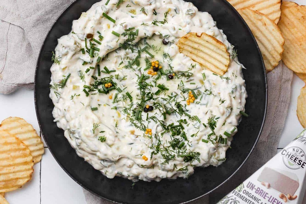 Bowl of dairy-free dill pickle dip topped with fresh herbs and served with potato chips next to a box of dairy-free brie cheese..