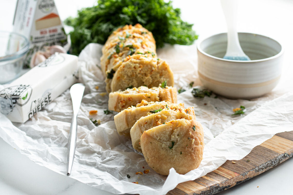 Baguette of dairy-Free cheesy garlic bread prepared and sliced. Served beside a bowl of melted dairy-free butter and a box of dairy-free gouda cheese.