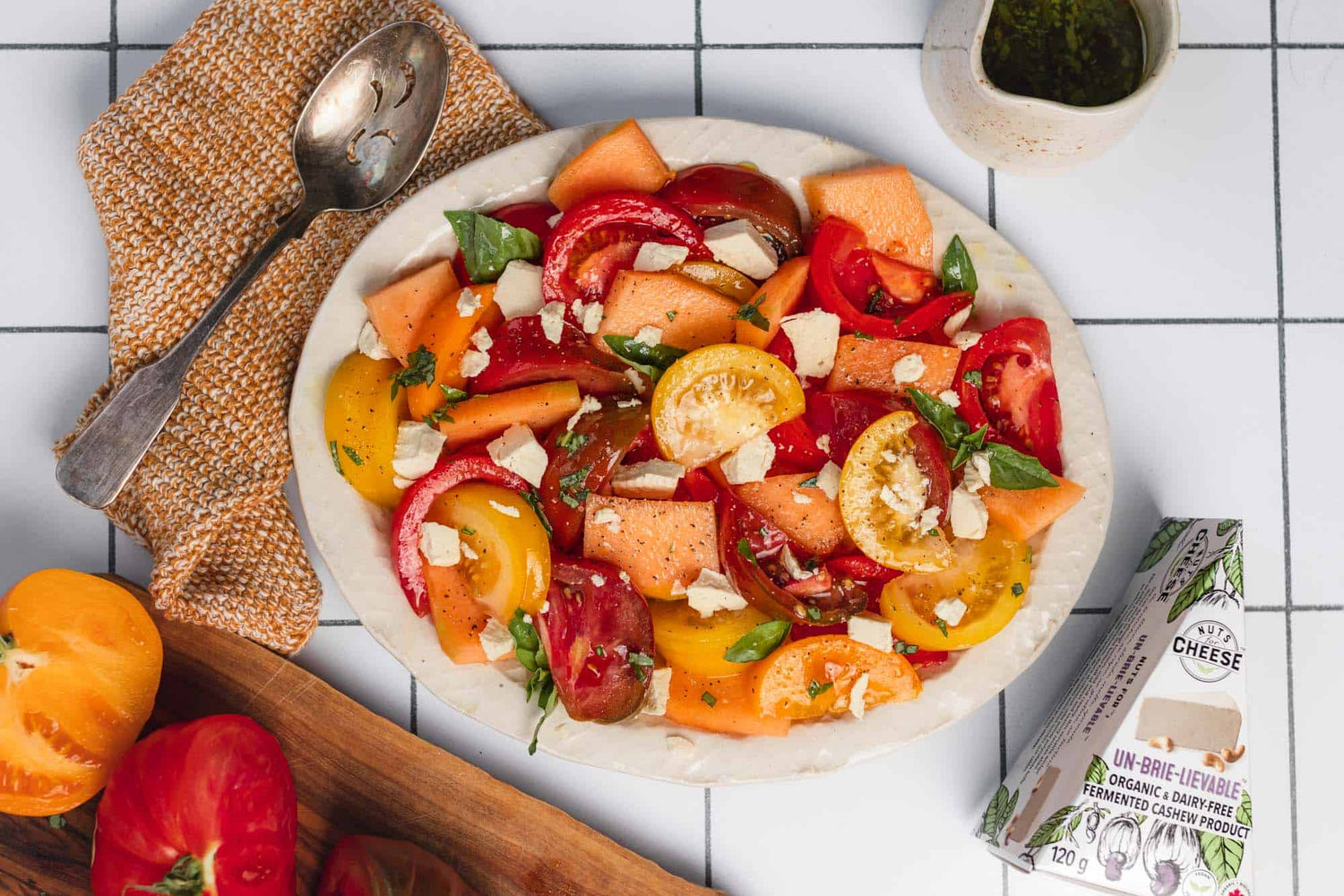 Prepared salad of sliced tomatoes, cantaloupe, and dairy free brie cheese, set on a napkin beside a box of dairy-free brie cheese and a serving spoon..