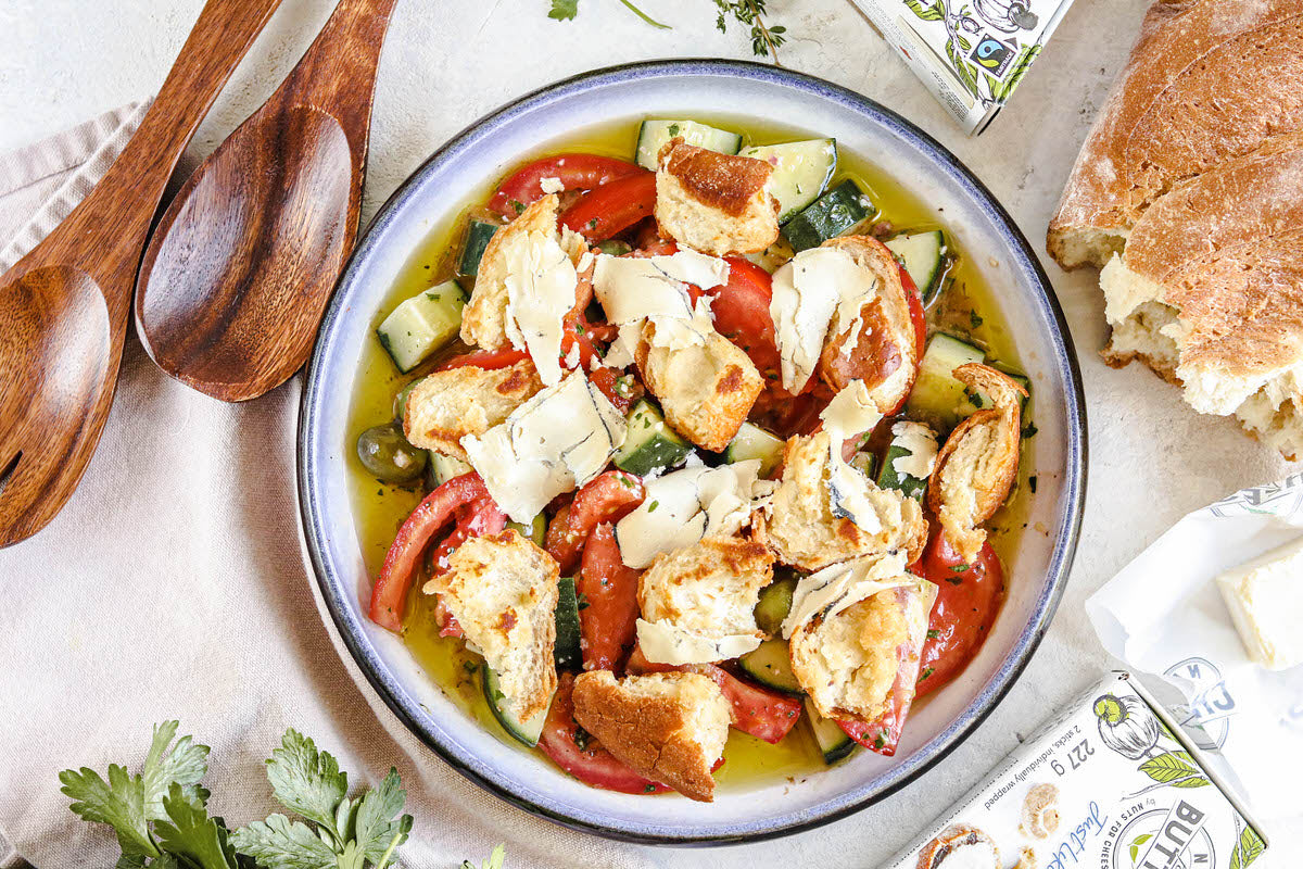Bowl of fresh panzanella salad featuring tomatoes, cucumbers, crispy bread, and pieces of dairy-free black garlic cheese. Served with a loaf of crusty bread.