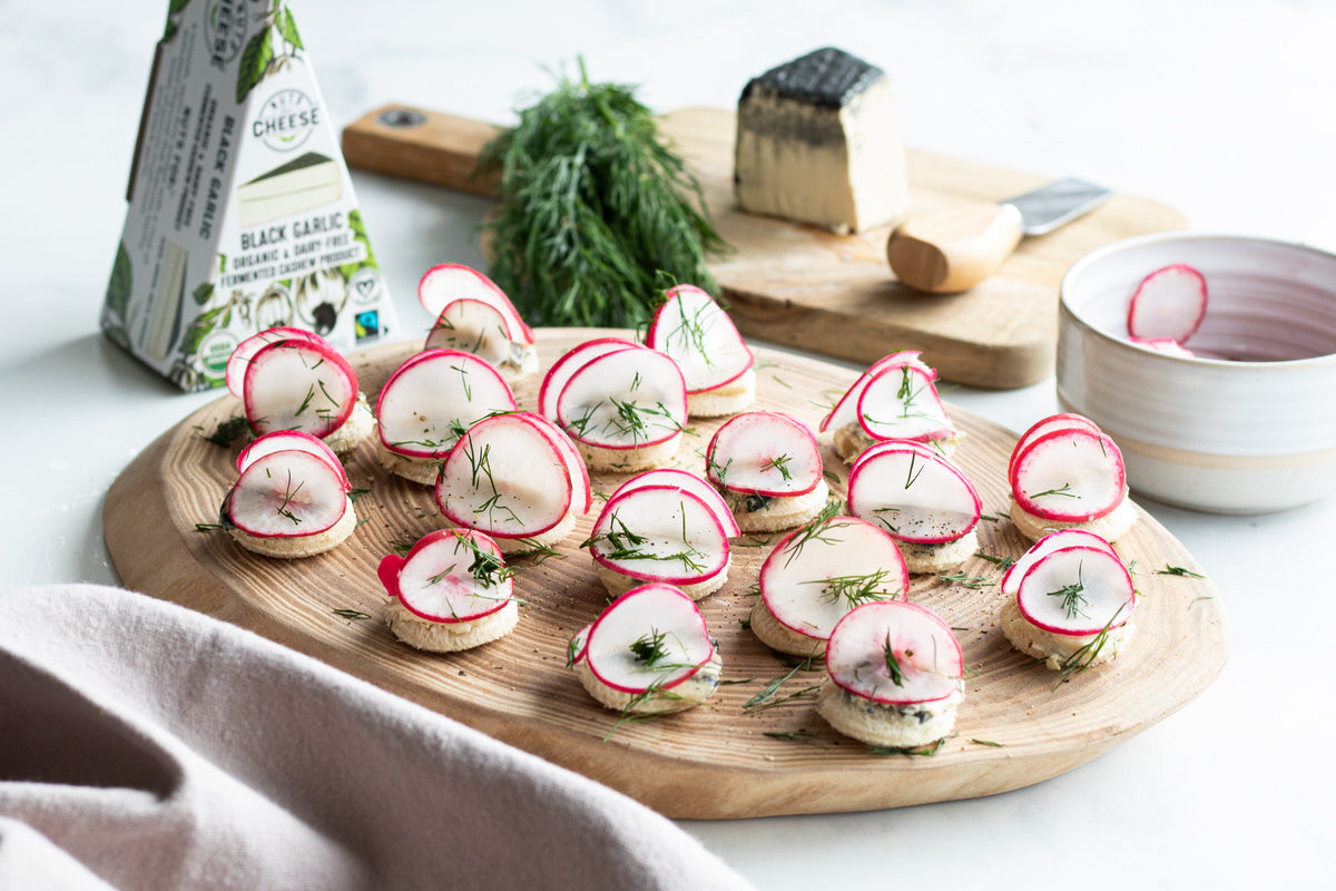 Wooden platter topped with radish canapes made from sliced bread, dairy-free cheese, sliced radishes and topped with fresh herbs. Served next to a box and sliced wedge of dairy-free black garlic cheese.