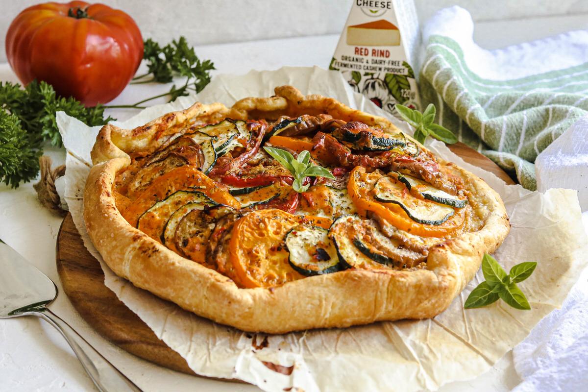 Baked galette in ratatouille-style featuring roasted tomatoes and zucchini and baked with a dairy-free gouda cheese. Served on a platter next to whole fresh tomatoes, fresh herbs, and a box of dairy-free smoky gouda cheese.