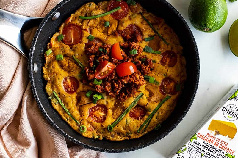 Skillet with a baked vegan frittata, made with dairy free chipotle cheddar cheese and topped with roasted tomatoes and vegan sausage next to a box of dairy free chiptotle cheddar cheese.