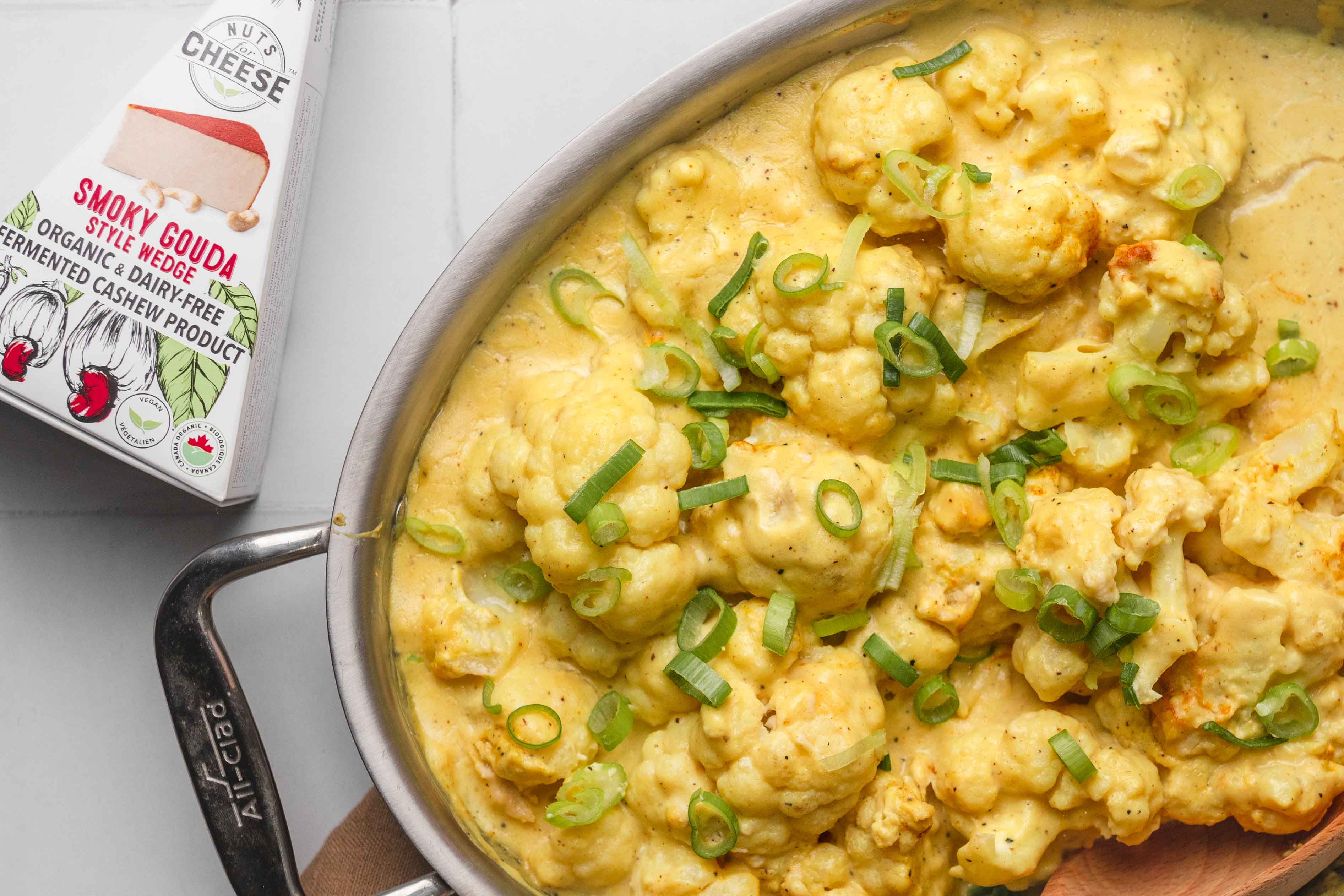 Pan filled with baked cauliflower in a dairy-free cheese sauce. Set next to a box of dairy-free gouda cheese.