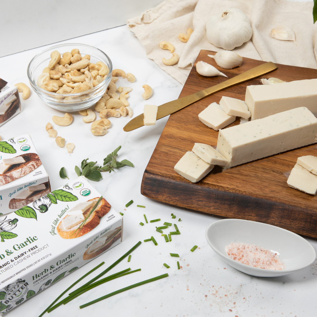 beurre à base de noix de cajou aux herbes et à l'ail assis sur une table avec un bol de noix de cajou, des quartiers d'ail et de beurre de noix de cajou