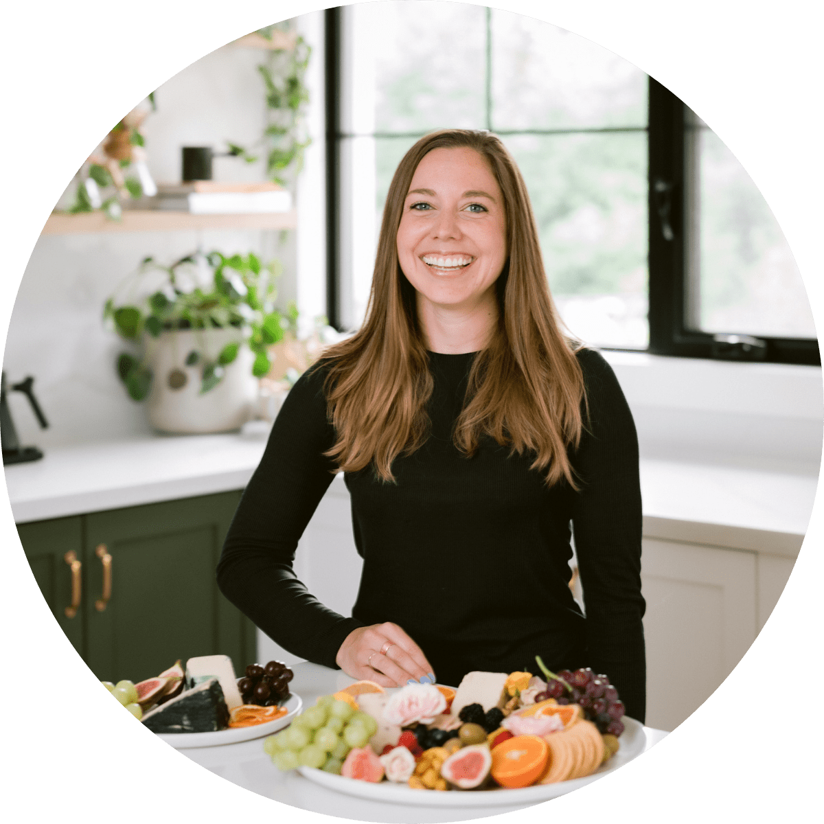 Nuts For Cheese Founder Margaret Coons smiling in a kitchen behind a plate of vegan cheese and fresh fruits.