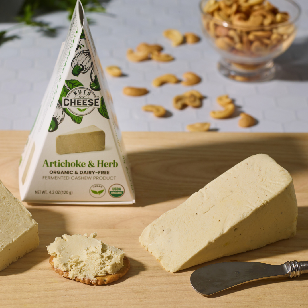 morceau de fromage de cajou aux artichauts et aux herbes et emballage sur une planche en bois avec des noix de cajou à l'arrière-plan