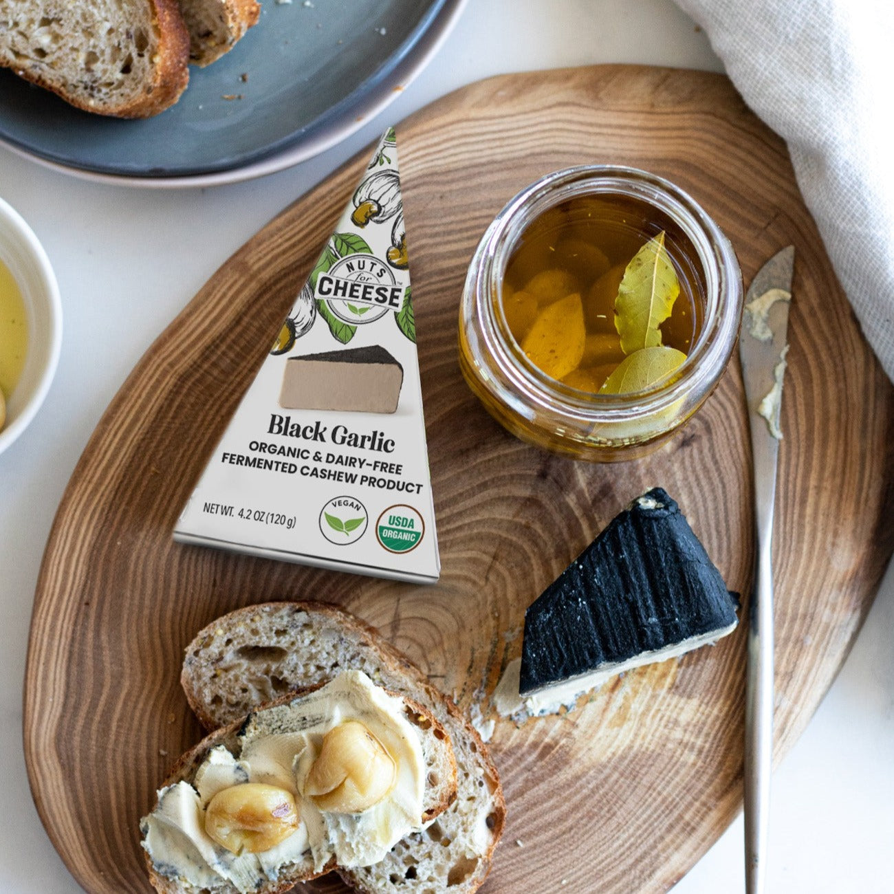 Nuts For Cheese dairy-free black garlic cheese wedge on a cutting board with bread slices and roasted garlic confit.