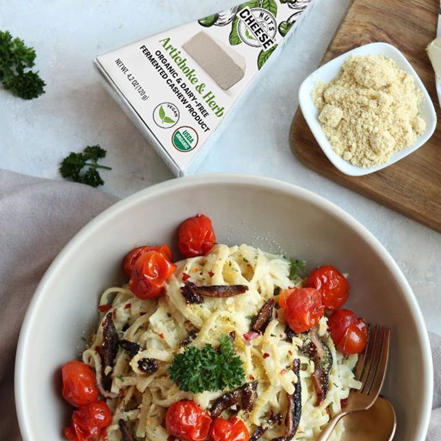 A plated spinach and herb carbonara with shiitake bacon dish with Nuts For Cheese™ Organic & dairy-free fermented Artichoke & Herb cheese wedge package