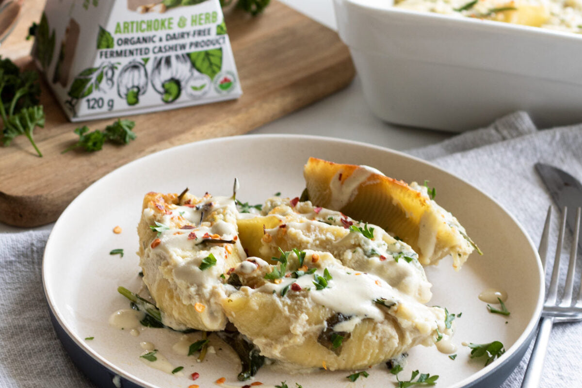 Plate with large pasta shells stuffed with dairy free cheese and topped with fresh herbs. Placed next to a box of dairy free artichoke & herb cheese.
