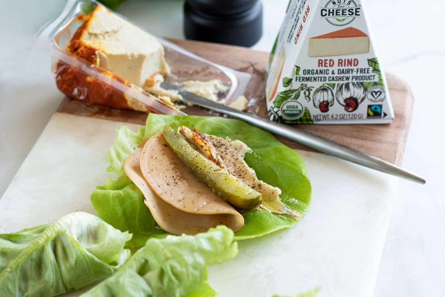 Lettuce wrap on a cutting board topped with vegan sliced turkey, dairy-free gouda cheese, and a pickle slice. Served next to a sliced wedge of dairy-free smoked gouda cheese.