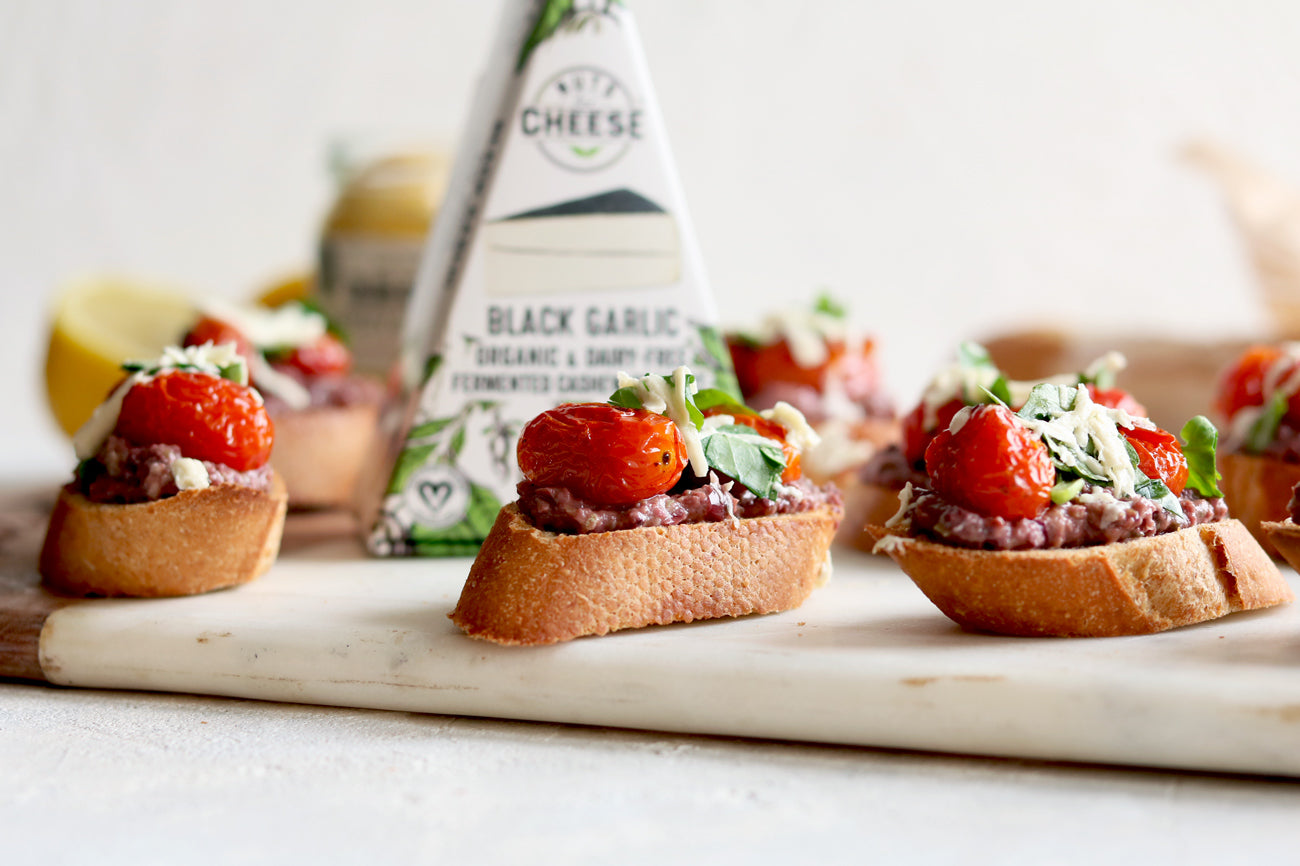 Three baguette slices topped with olive tapenade and dairy-free black garlic cheese, roasted cherry tomatoes, and fresh herbs. Served beside a box of dairy-free black garlic cheese.