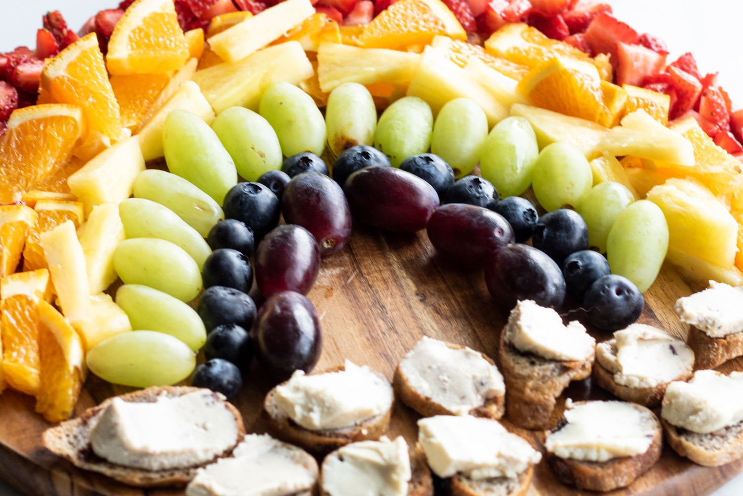 Platter of fruits arranged in a rainbow shape by color. Dairy-free cheese spread on baguette slices form a cloud shape at the base.