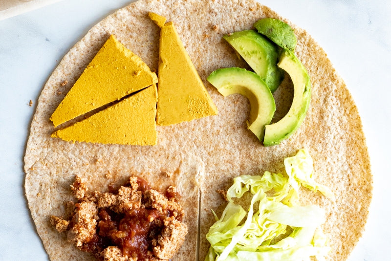 Open tortilla shell topped with one ingredient in each quadrant: dairy-free chipotle cheddar cheese, avocado, shredded lettuce, cooked vegan sausage.