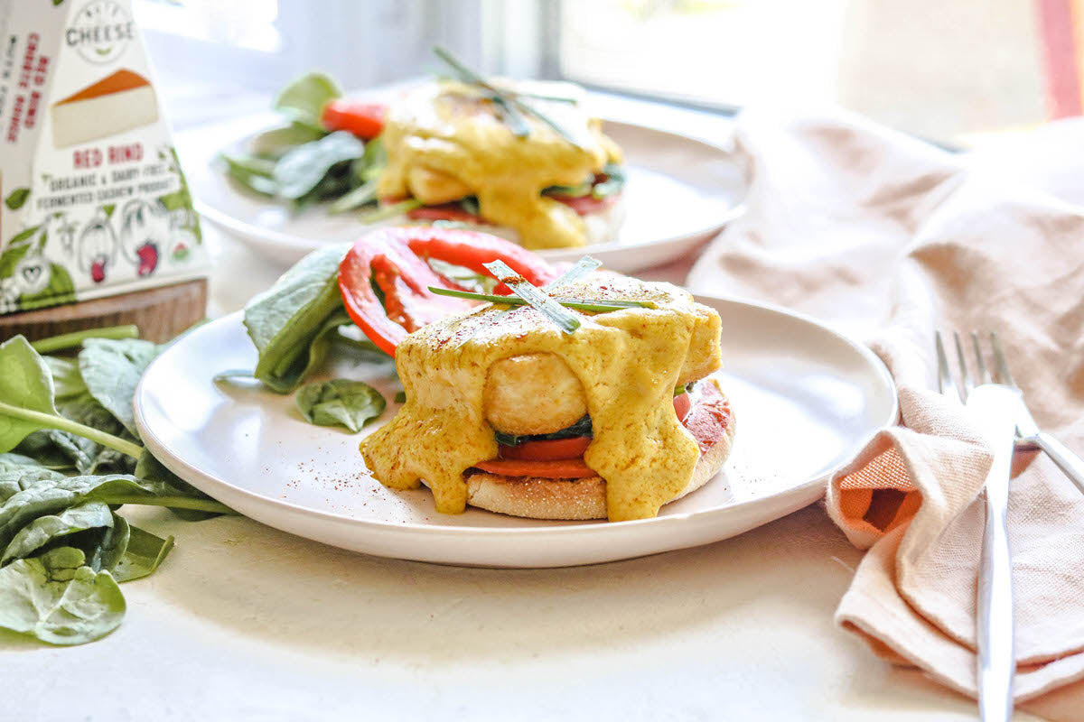 A plate with English muffins prepared with vegan eggs and a dairy-free hollandaise sauce. Served with fresh green salad next to a box of dairy-free smoky gouda cheese.