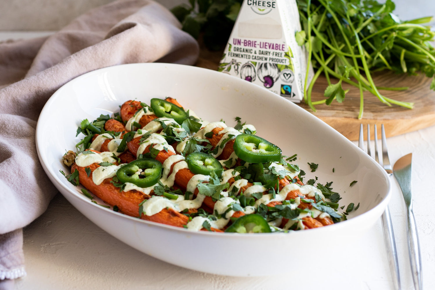 Large serving bowl filled with roasted carrots, topped with a dairy-free cheese sauce, sliced jalapenos, and fresh cilantro. Served next to a box of dairy-free brie cheese and bunch of fresh cilantro.
