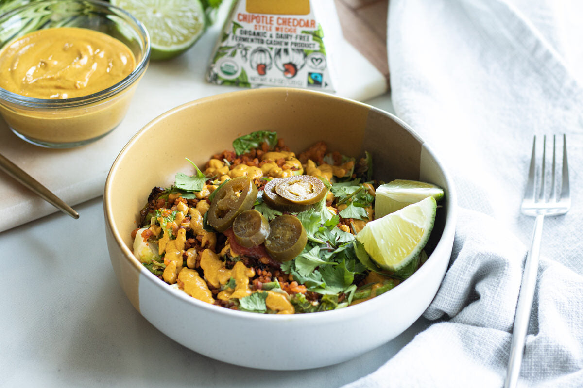 A bowl of brussels sprout nachos topped with dairy-free chipotle cheddar cheese sauce, pickled jalapenos, and limes. Served with a side of dairy-free cheese sauce and next to a box of dairy-free chipotle cheddar cheese.