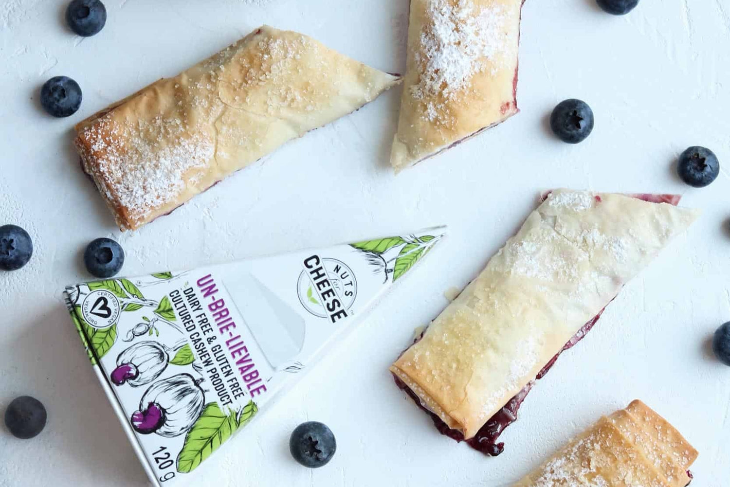 Baked phyllo rolls stuffed with dairy-free brie cheese and a balsamic blueberry compote. Fresh blueberries are sprinkled around and a box of dairy-free brie cheese is next to the rolls.