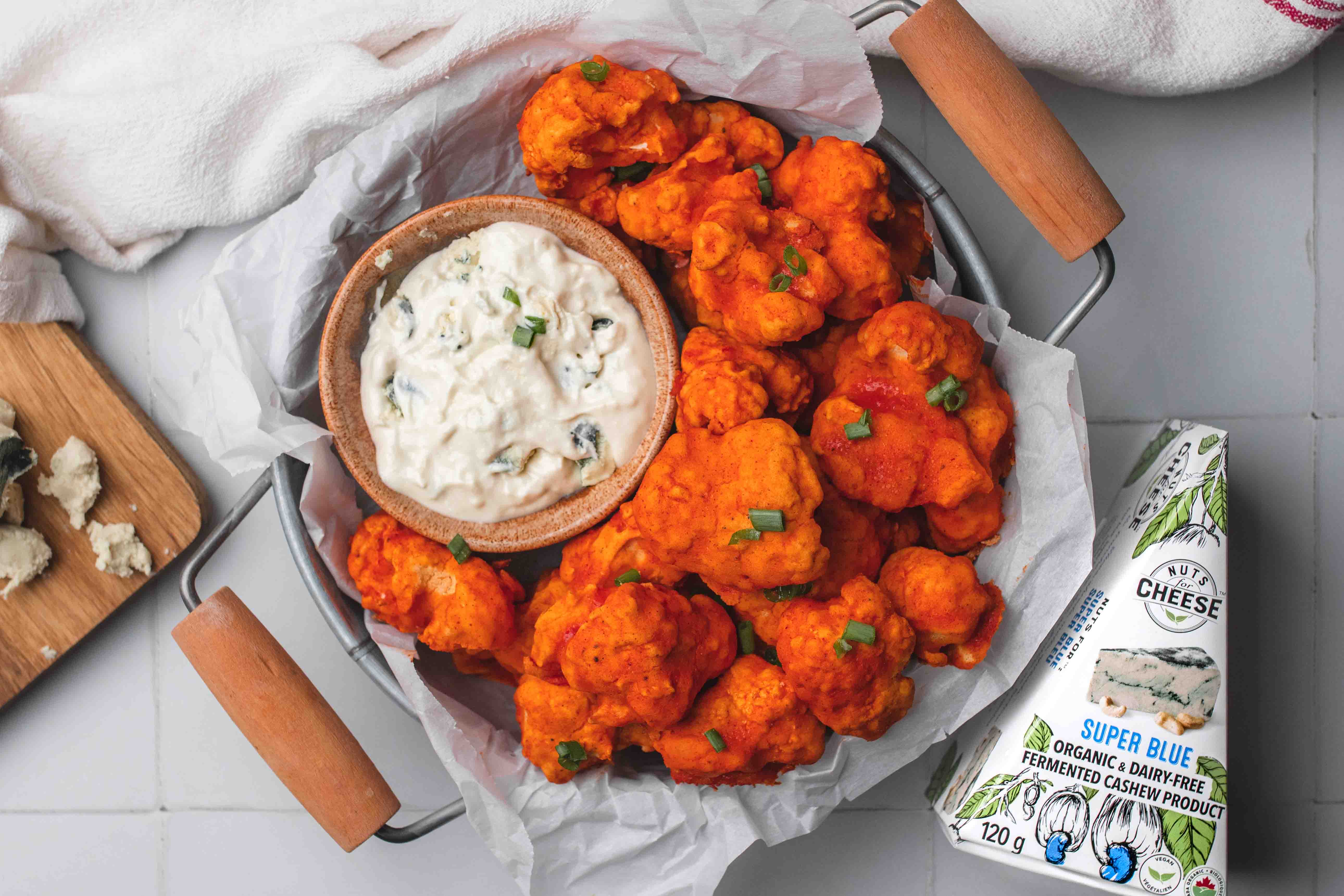 Bowl of buffalo-style cauliflower served with a side of dairy-free blue cheese dip beside a box of dairy-free blue cheese.