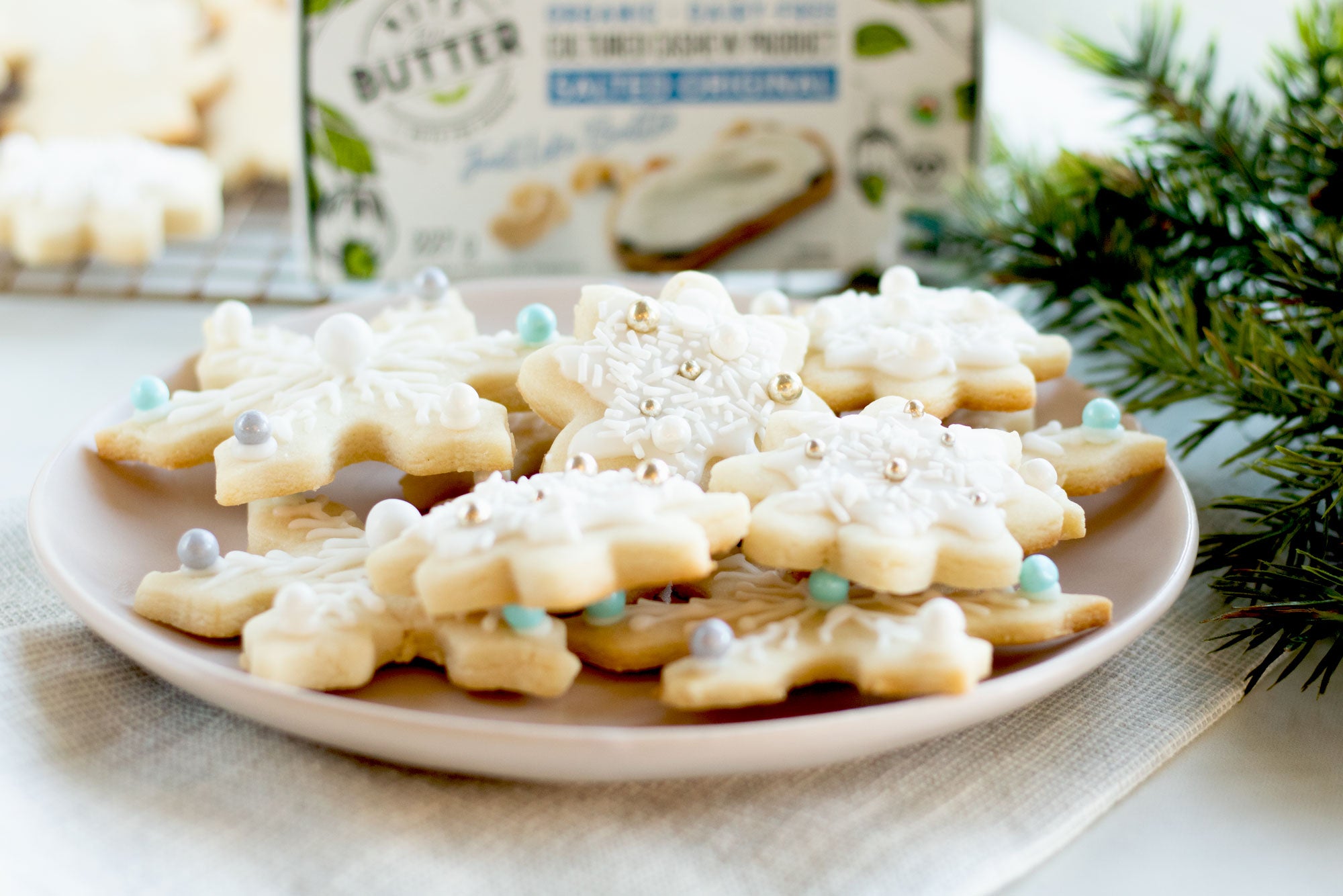Plate of snowflake-shaped vegan butter sugar cookies topped with white icing and sprinkles. In the background is a box of dairy-free butter and more cookies on a cooling rack.