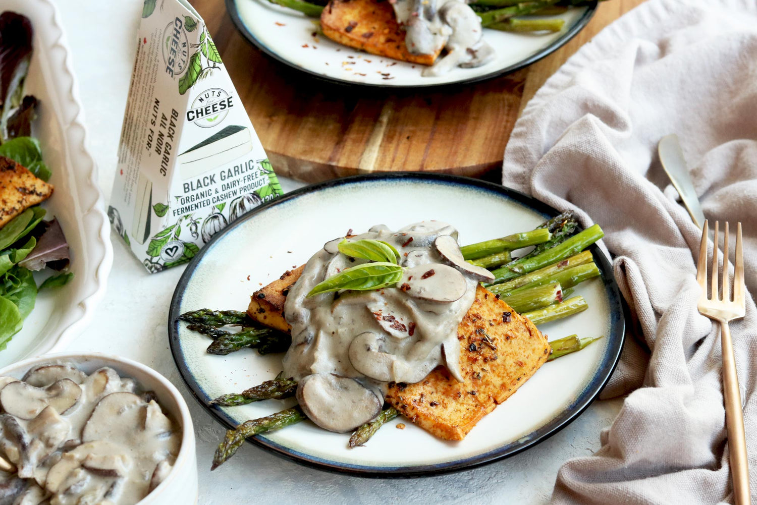 Plate of cooked asparagus topped with a smoked tofu steak and drizzled with a dairy-free black garlic mushroom gravy. Served next to a box of dairy-free black garlic cheese and an extra bowl of vegan mushroom gravy.