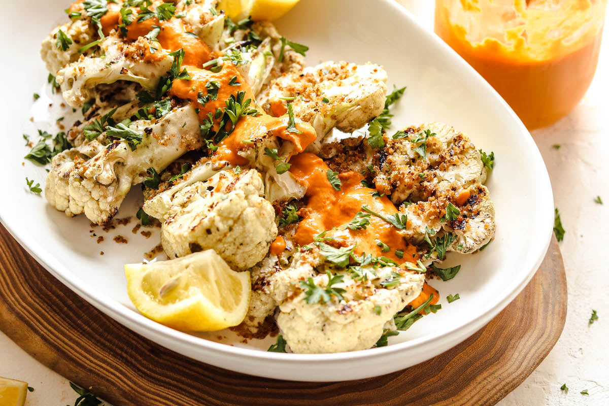 A plate of grilled cauliflower drizzled with a dairy-free cheesy red pepper sauce and fresh herbs. 