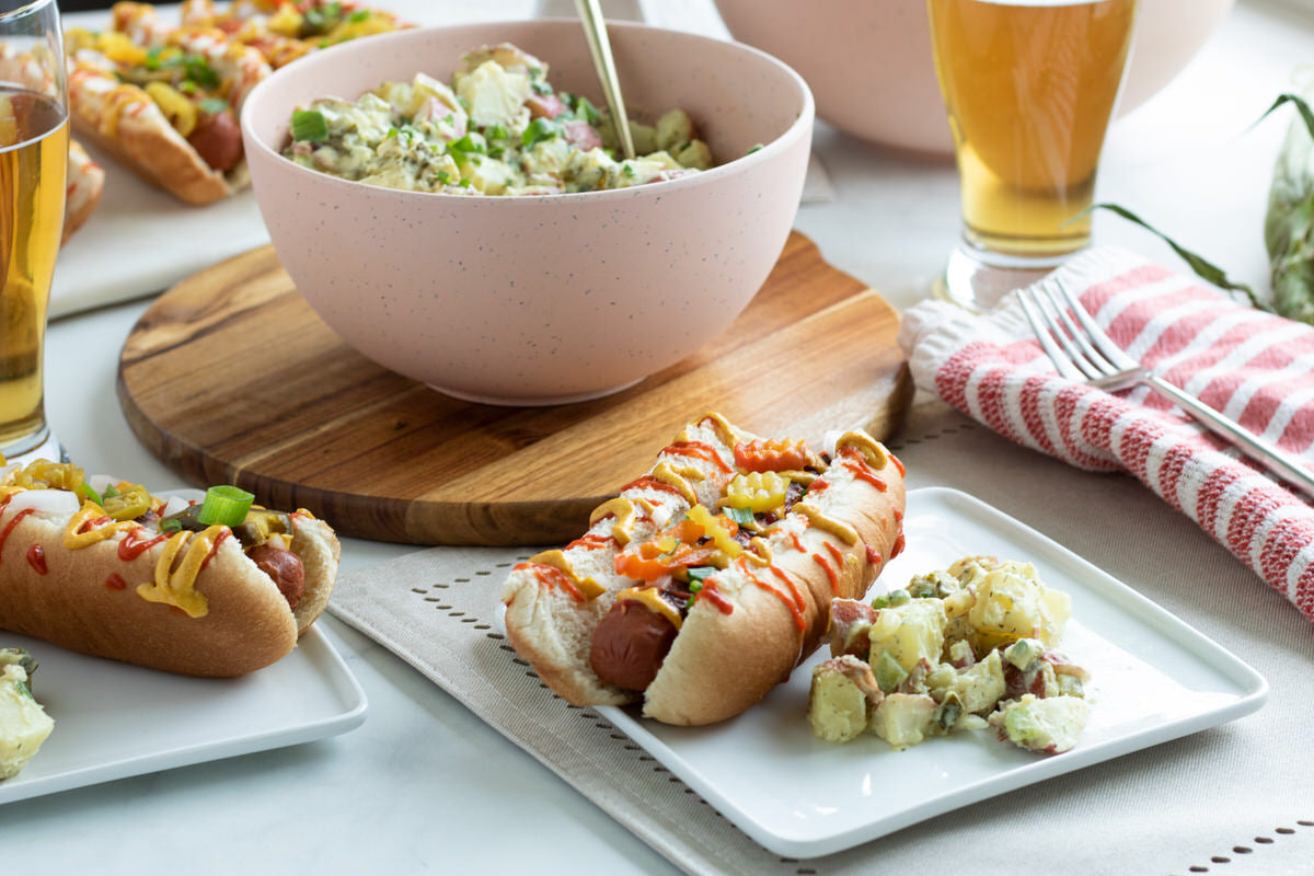 Two plates with prepared vegan hot dogs topped with condiments and served next to cheesy vegan potato salad with a large bowl of vegan potato salad in the background.