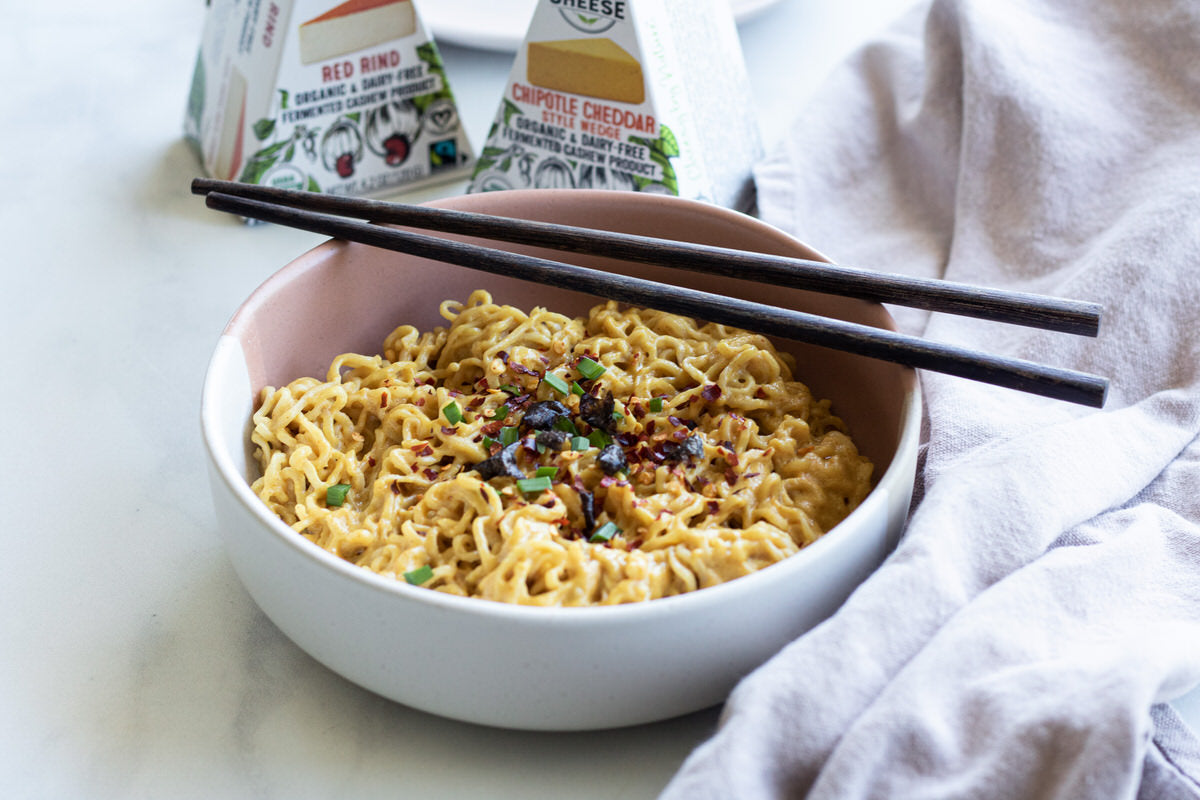 Bowl of cheesy vegan ramen with wood chopsticks. Served beside two boxes of dairy-free cheese.