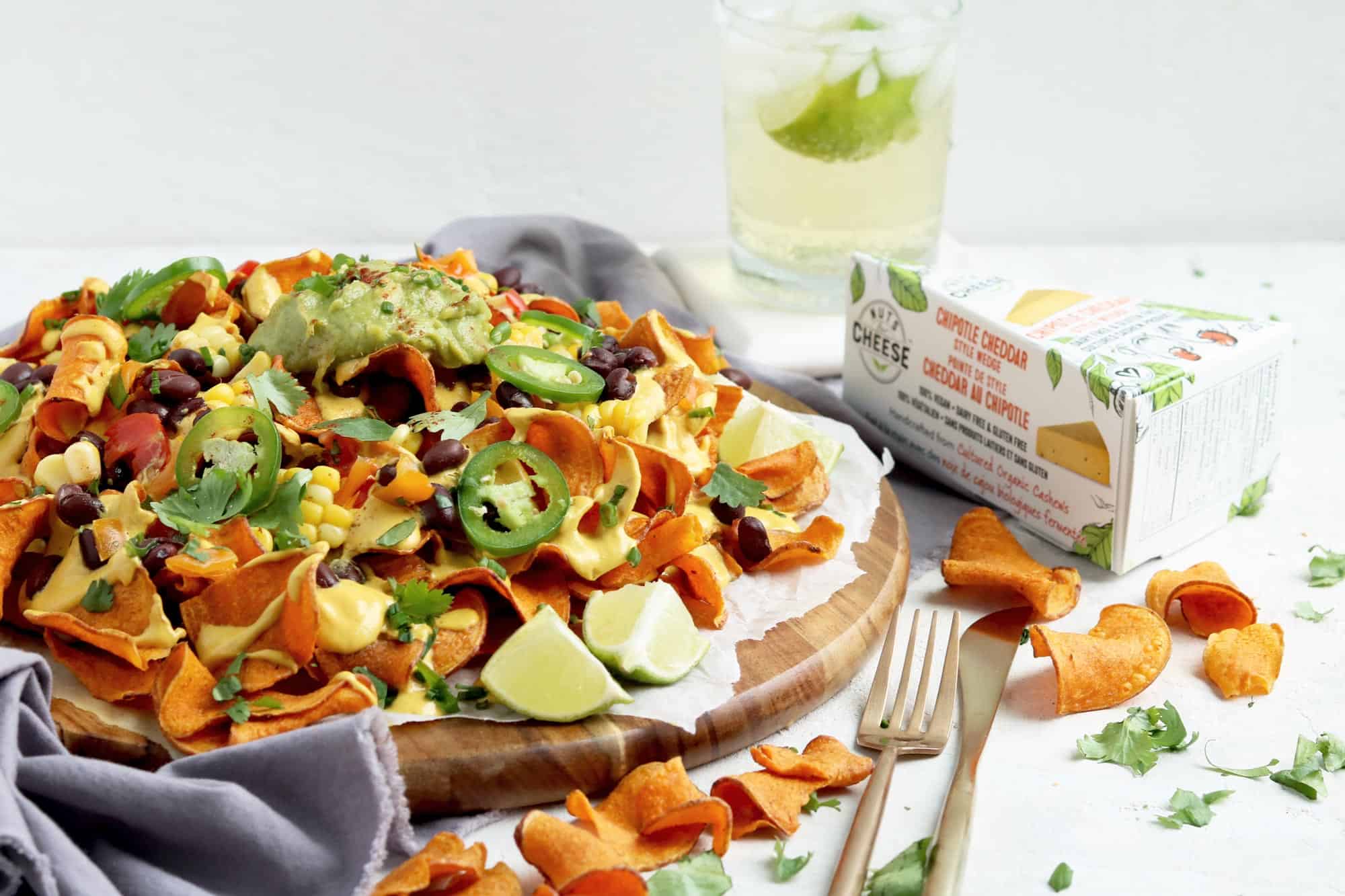 Plate of sweet potato nachos loaded with dairy-free chipotle cheese sauce, corn, black beans, jalapenos and guacamole. Served beside a box of dairy-free chipotle cheddar cheese.