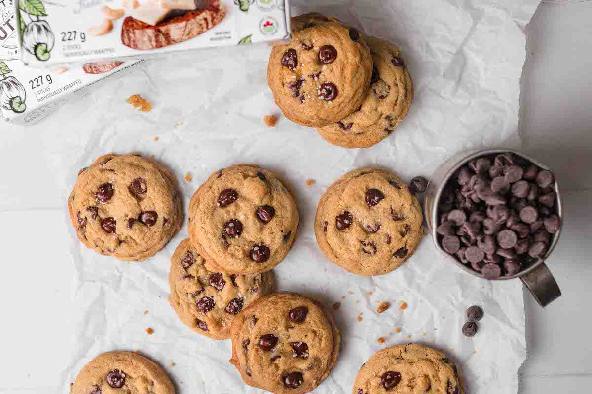 Dairy-free chocolate chip cookies scattered about next to boxes of dairy-free butter and a cup of chocolate chips.