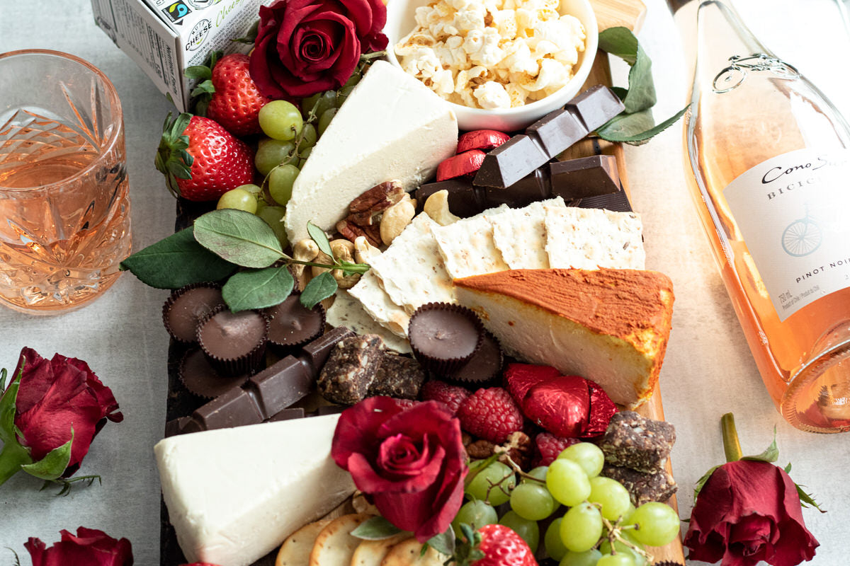 A dairy-free dessert cheeseboard featuring wedges of dairy-free brie, dairy-free gouda, grapes, strawberries, vegan chocolate, and red roses.