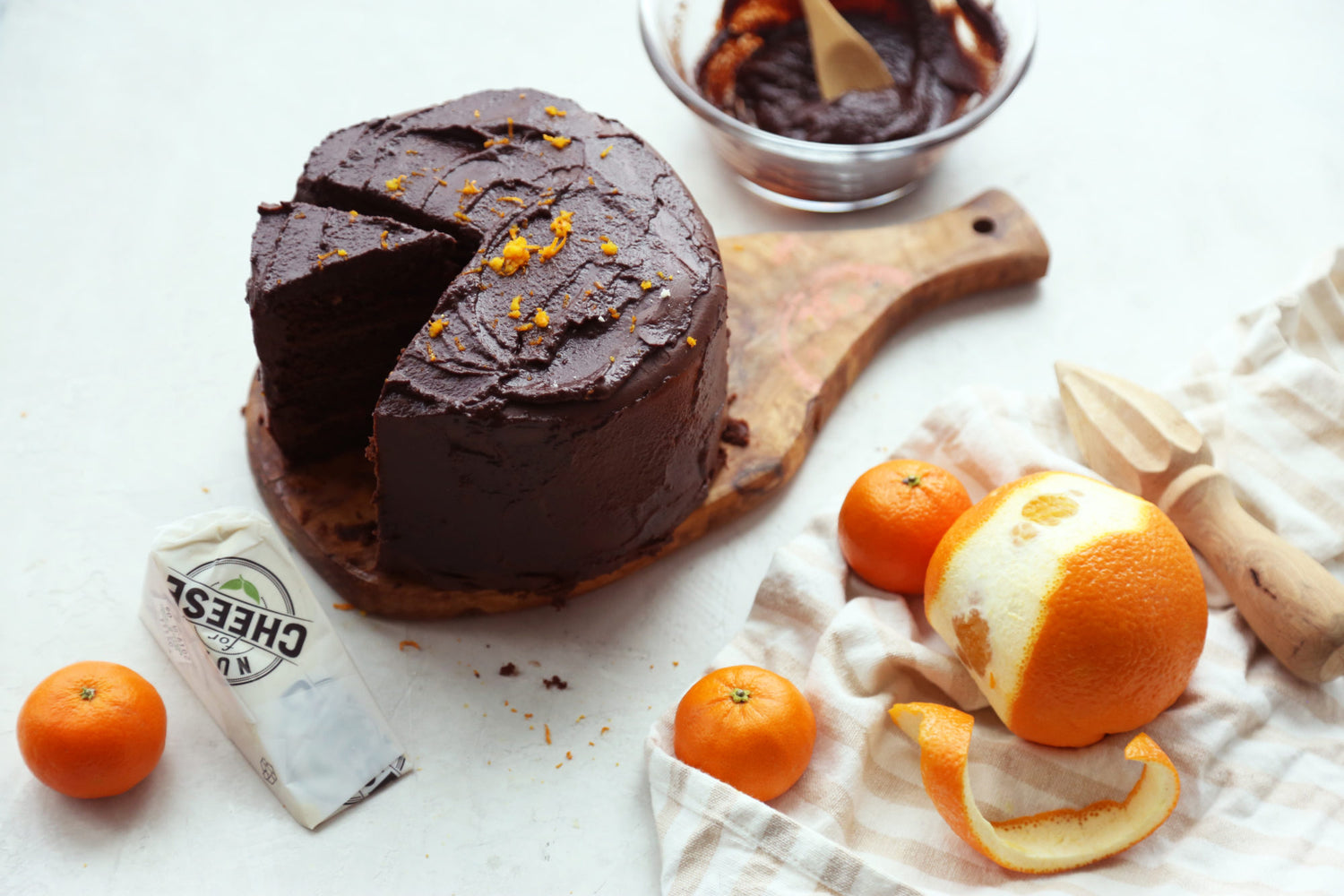 Double layer chocolate cake with a slice cut out, made with hints of orange and dairy free cheese sits on a cutting board next to fresh oranges and a wedge of dairy-free cheese.