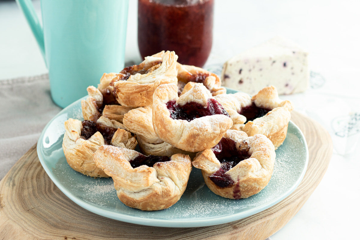 A plate of baked puff pastries stuffed with dairy-free cheese and raspberry jam sit on a plate beside a wedge of dairy-free cranberry pink peppercorn cheese.