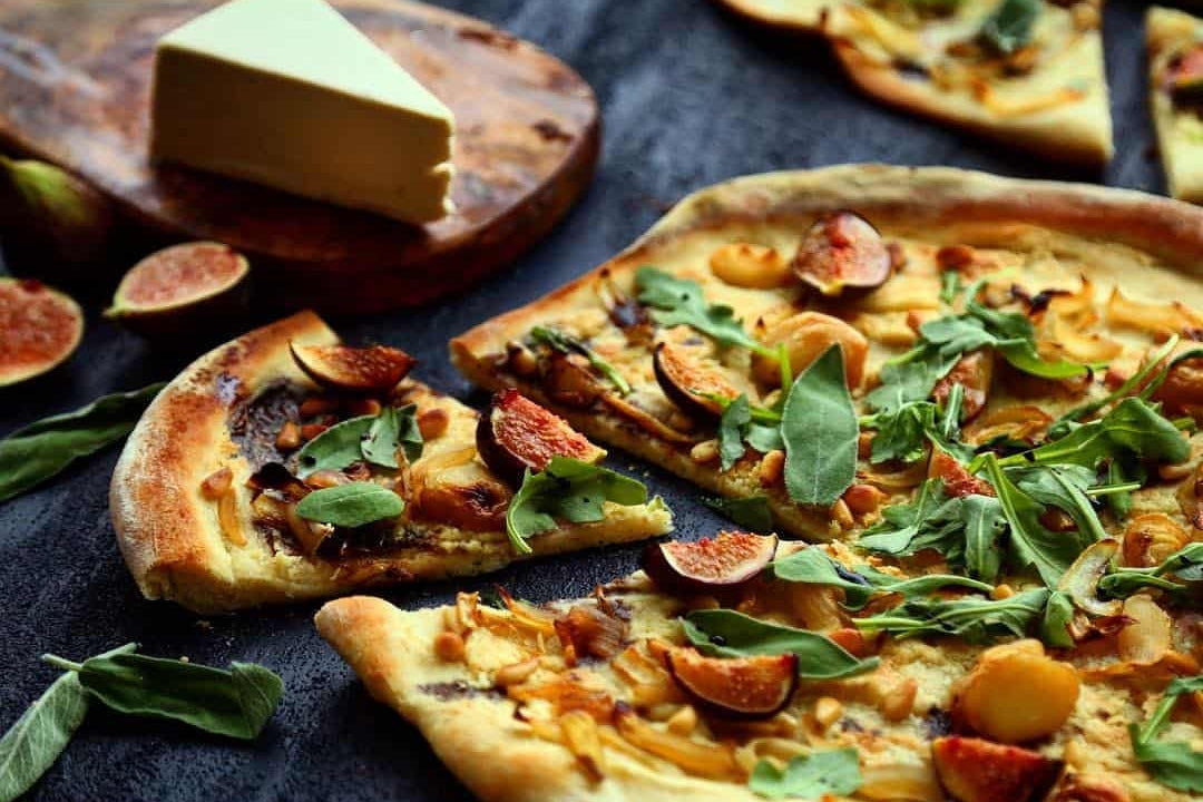 Baked pizza topped with fresh figs, roasted garlic, carmelized onions sits beside a wedge of dairy-free brie cheese.