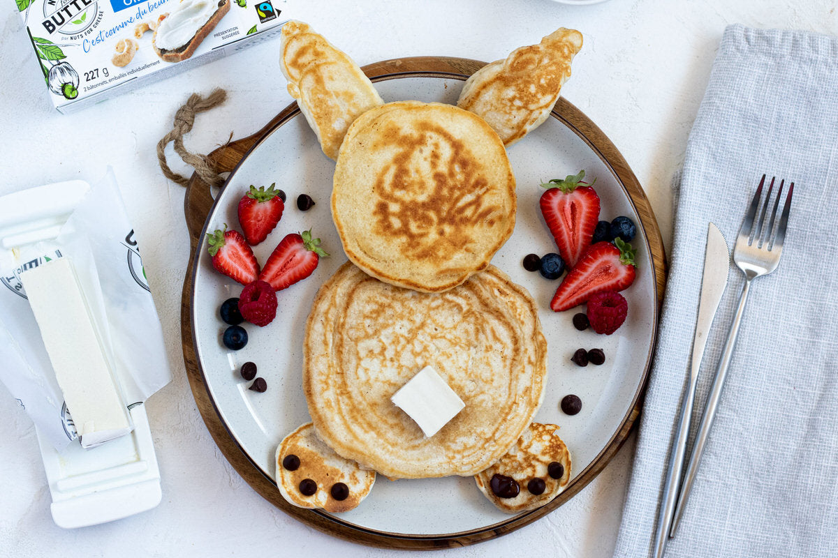 Plate of vegan pancakes arranged in the shape of a bunny, served with fresh berries and a pat of dairy-free butter. Served next to a stick of dairy-free butter, napkin, and fork.