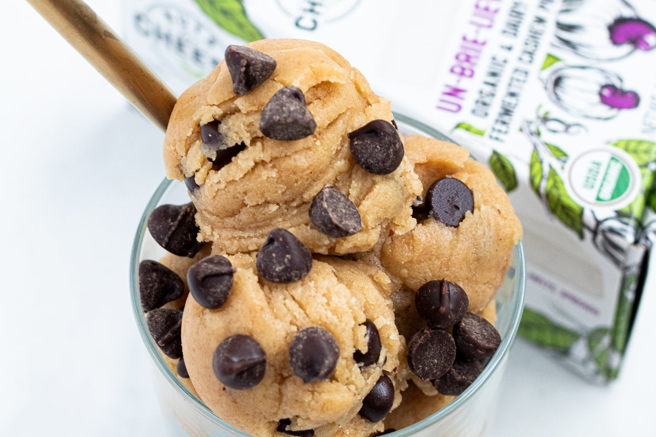 Three scoops of vegan edible cookie dough with chocolate chips are served in a cup beside a box of dairy-free brie cheese.