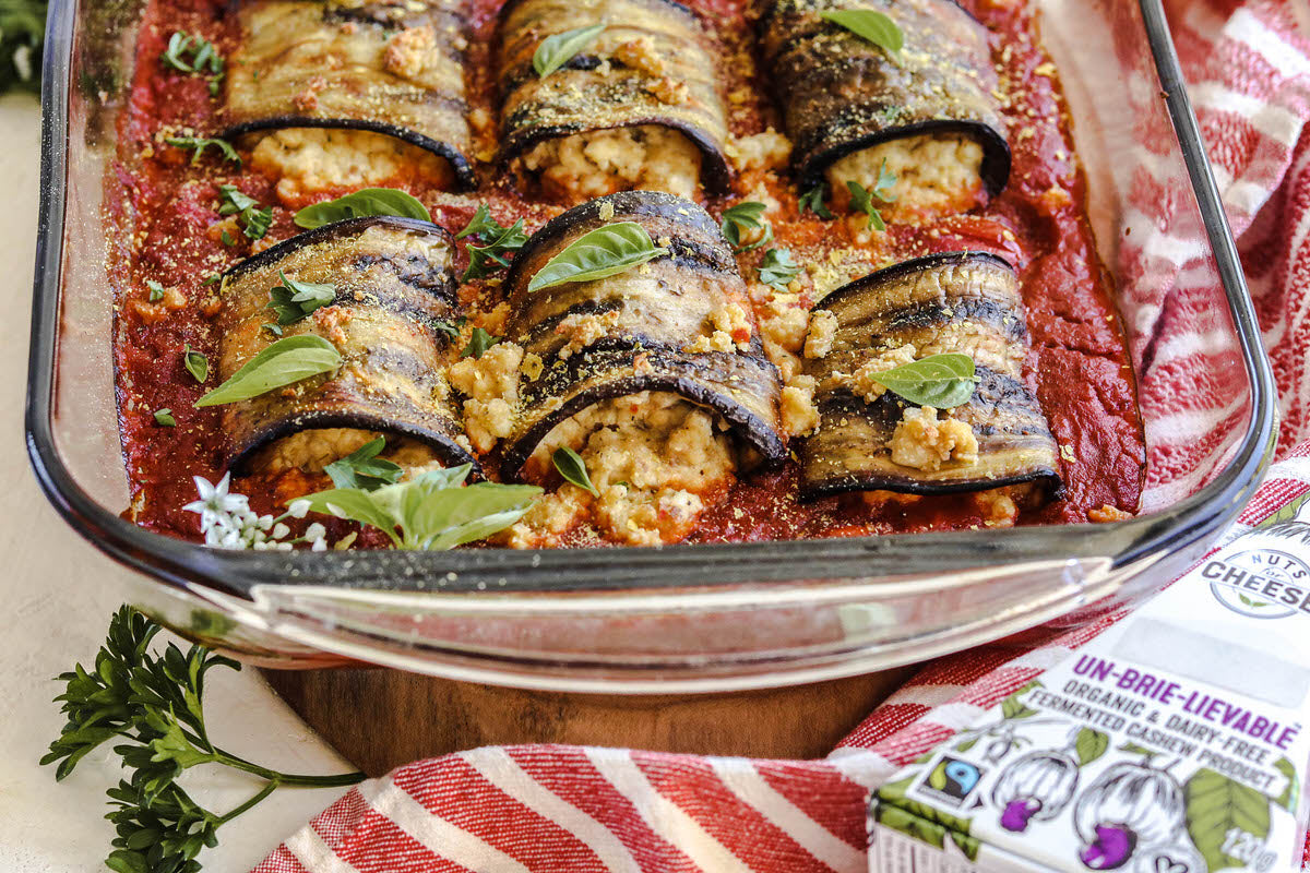 Baking dish filled with prepared eggplant involtini stuffed with dairy-free brie cheese served next to a box of dairy-free brie cheese.