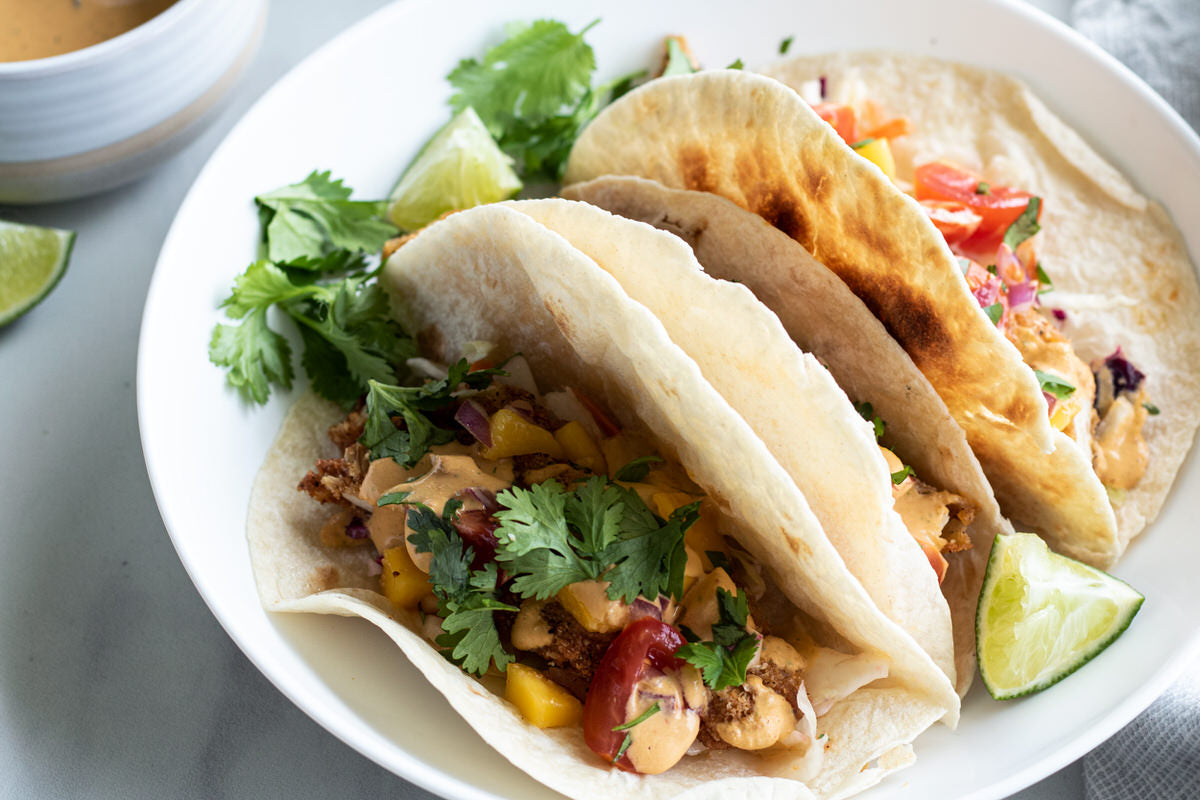 Two vegan fish tacos sit on a plate. The tacos are topped with a dairy-free chipotle lime cheese sauce and fresh cilantro.